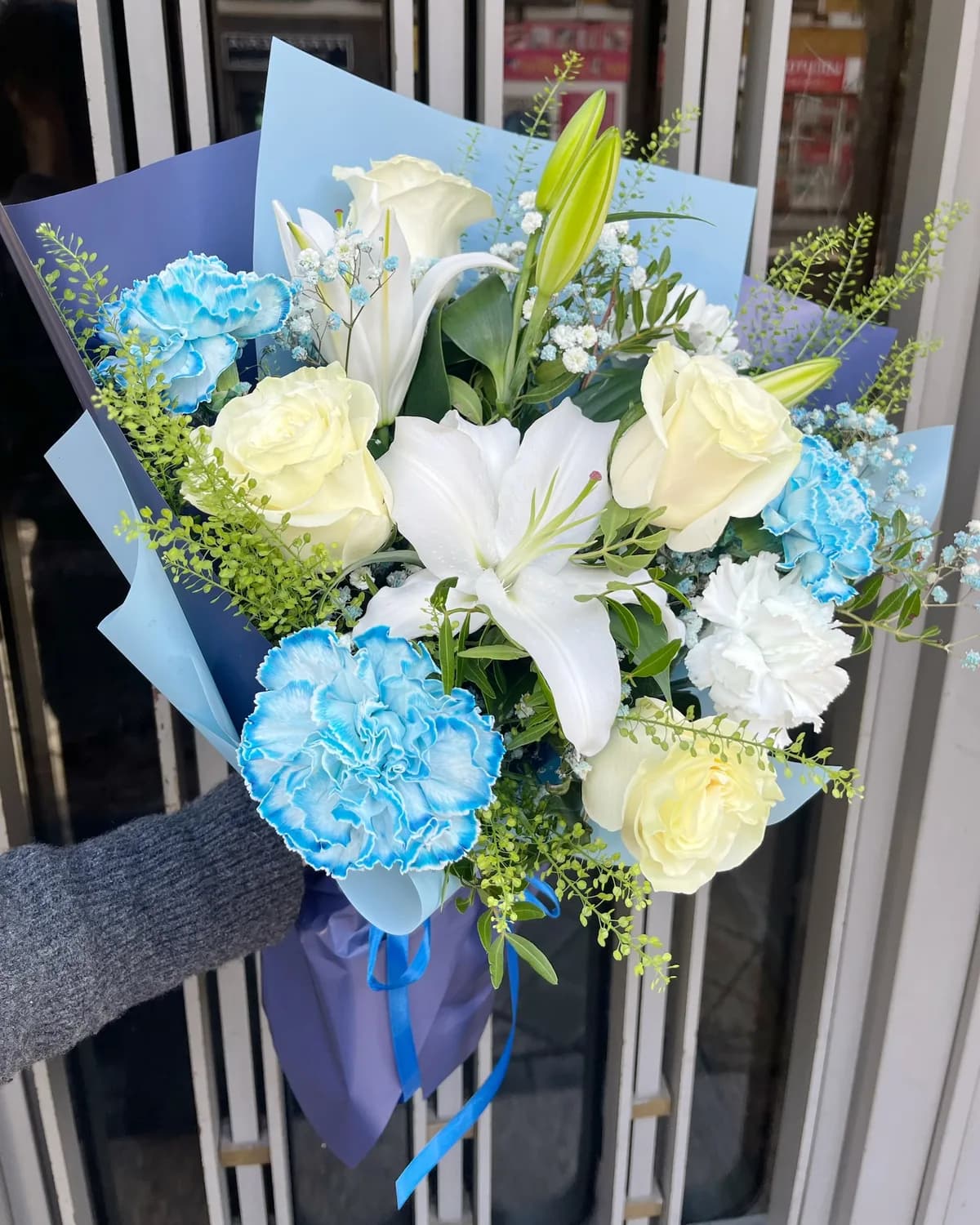 Blue and white bouquet of white roses, lilies and blue carnations in light and navy blue paper with a satin ribbon